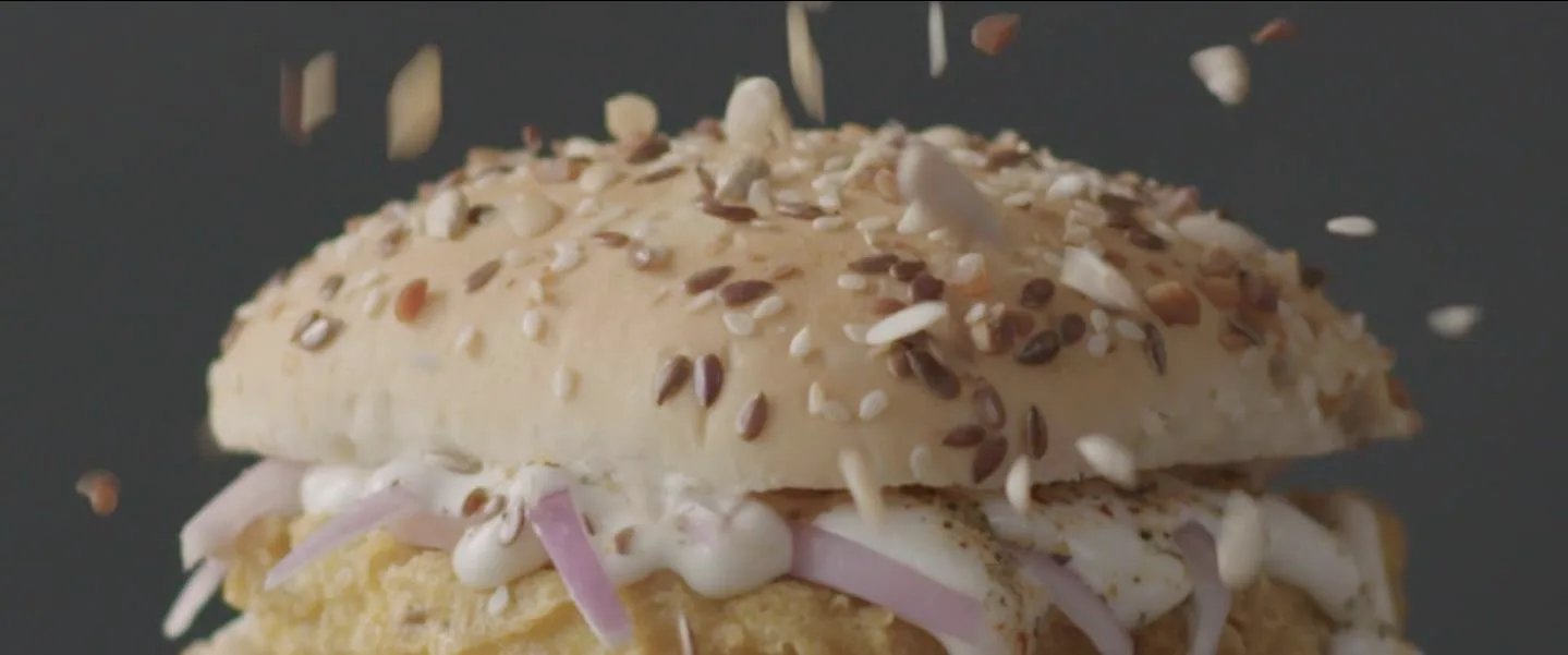 Cinematic food-commercial still: extreme close-up of a multigrain burger bun crowned with flax, sesame and sunflower seeds mid-air-drop against a charcoal-grey seamless backdrop, seeds and nut slivers frozen in flight around it, with mayonnaise, sliced red onion and a golden fried patty visible under the bun.