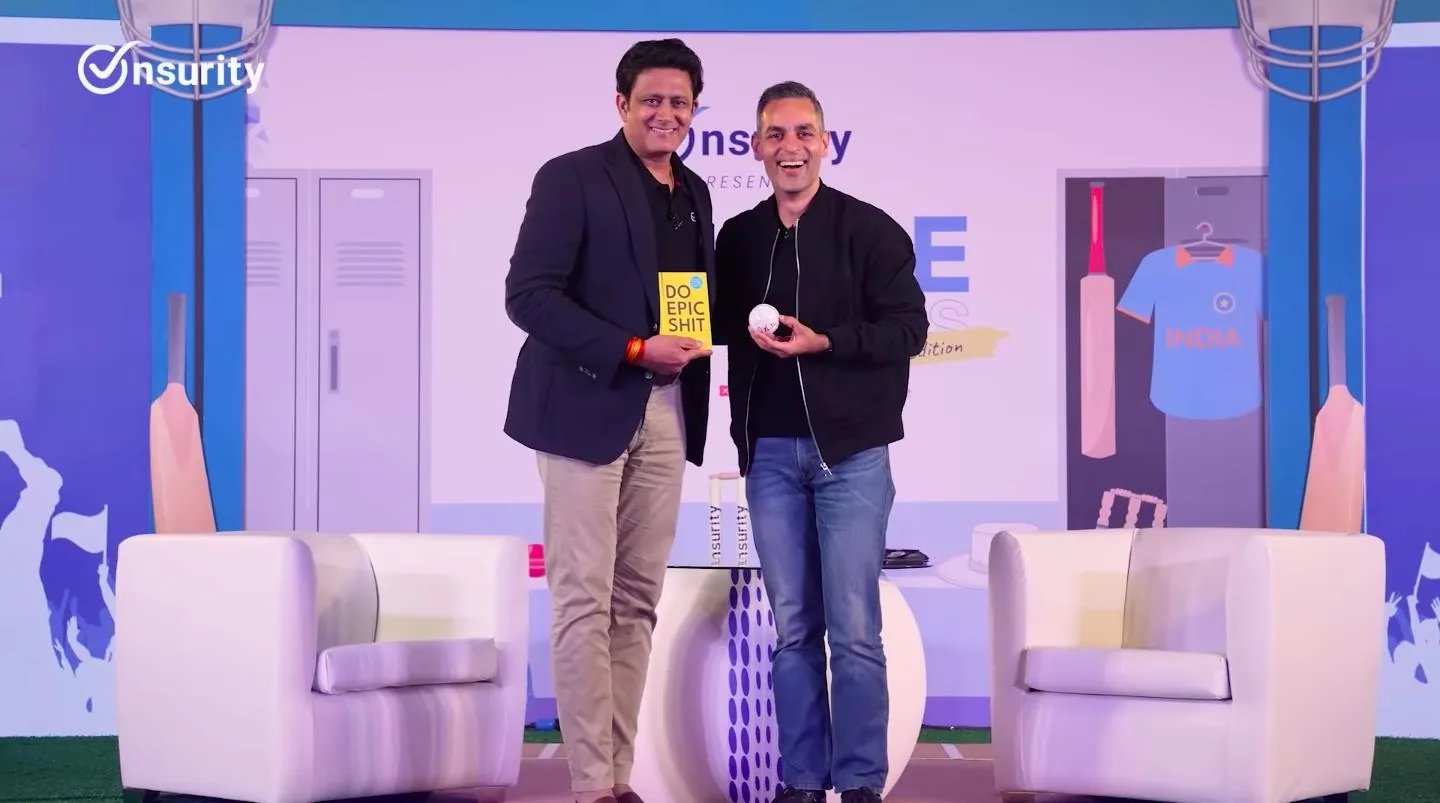 Two male speakers standing centre-stage at the Onsurity x Keka influencer event; the set is built as a cricket dressing room with grey lockers, a hanging India jersey, cricket bats, stumps and a wicket-keeper glove backdrop; one speaker holds a yellow 'Do Epic Shit' book, the other a white cricket ball; two cream armchairs and a branded round table on astroturf flooring, large Onsurity logo top-left of the backdrop