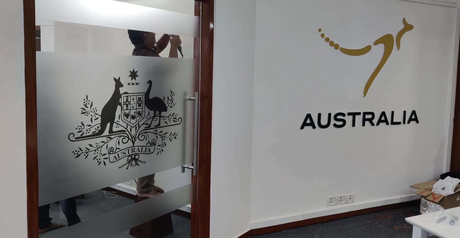 Office entrance with a frosted-vinyl glass door carrying the Australian Commonwealth Coat of Arms (kangaroo and emu flanking the shield) and, on the adjacent white wall, the gold Australia Nation Brand kangaroo mark above 'AUSTRALIA' in dark grey dimensional lettering