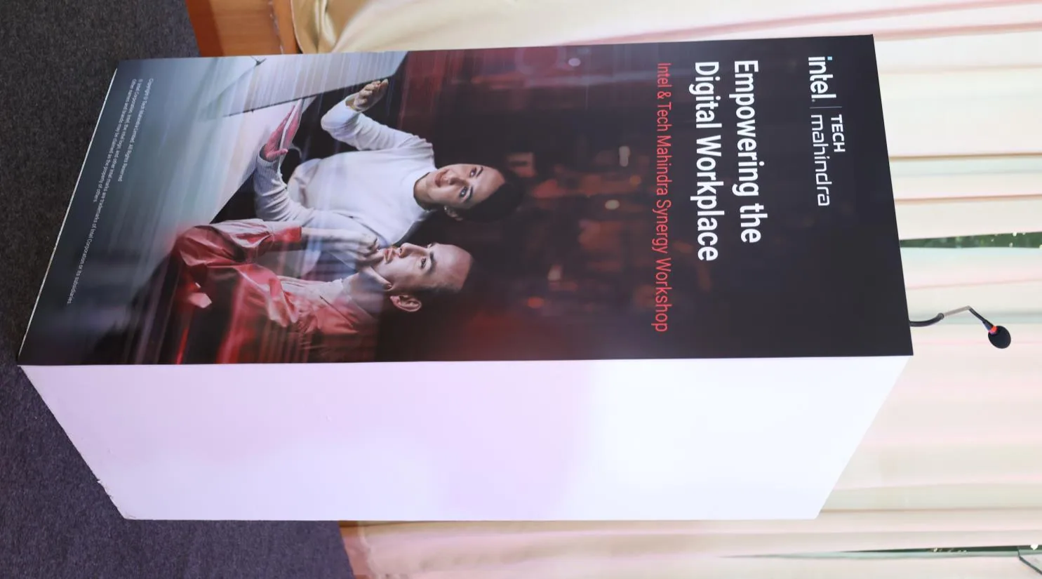 Event photo of a white podium with a black branded front panel carrying the Intel/Tech Mahindra 'Empowering the Digital Workplace' artwork, gooseneck microphone attached, cream drape curtain and grey carpet behind