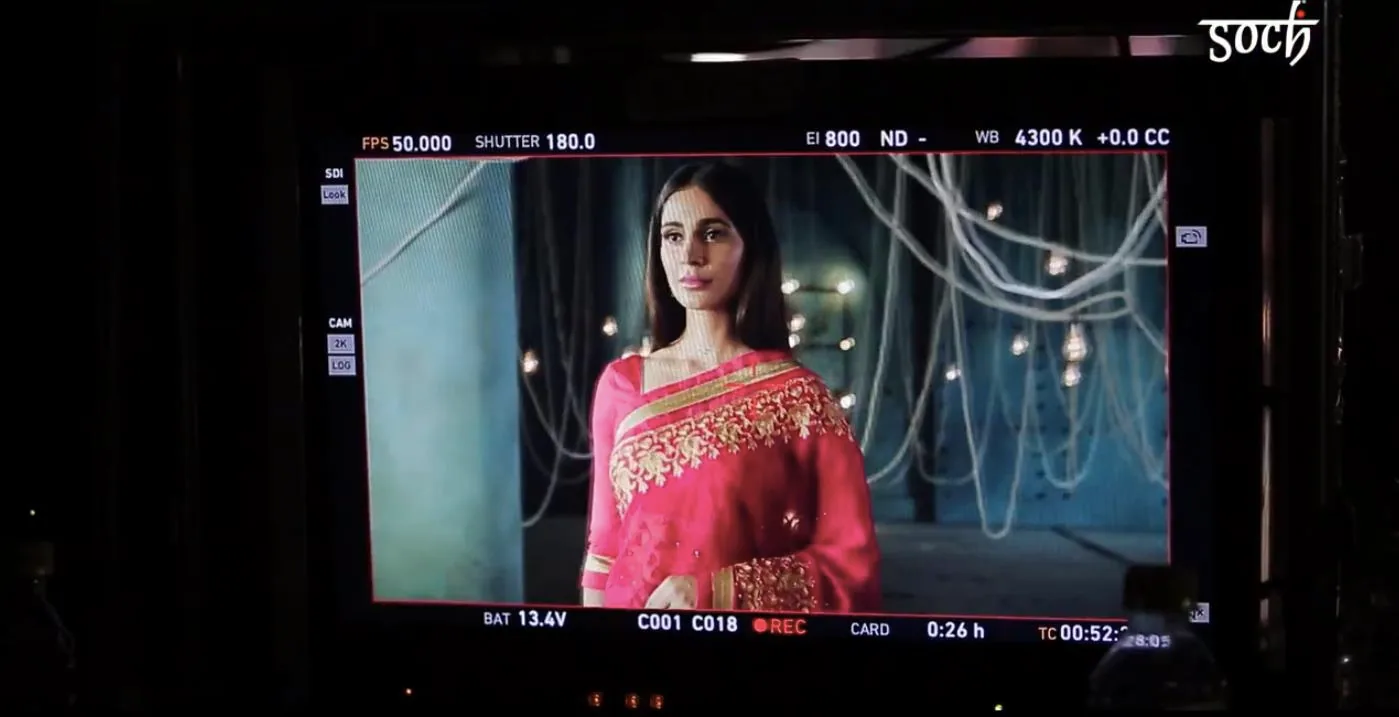 Behind-the-scenes still of the on-set director's monitor in a darkened room, its screen showing the live recording frame of a model in a bright pink gold-embroidered saree against a teal wall hung with bulb strings; monitor overlay reads FPS 50.000, Shutter 180.0, EI 800, WB 4300 K, C001 C018 REC, TC 00:52; 'Soch' logo watermarked top right